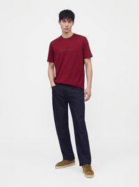 Hombre con una camiseta roja de manga corta, pantalones azul oscuro y zapatillas marrones, de pie con una mano en el bolsillo sobre un fondo blanco.
