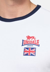 Balta trumpa rankovėmis marškinėliai su tamsiai mėlynu įmigtu apykakle; ant krūtinės yra raudonas "Lonsdale London" logotipas ir Jungtinės Karalystės vėliavos grafika.