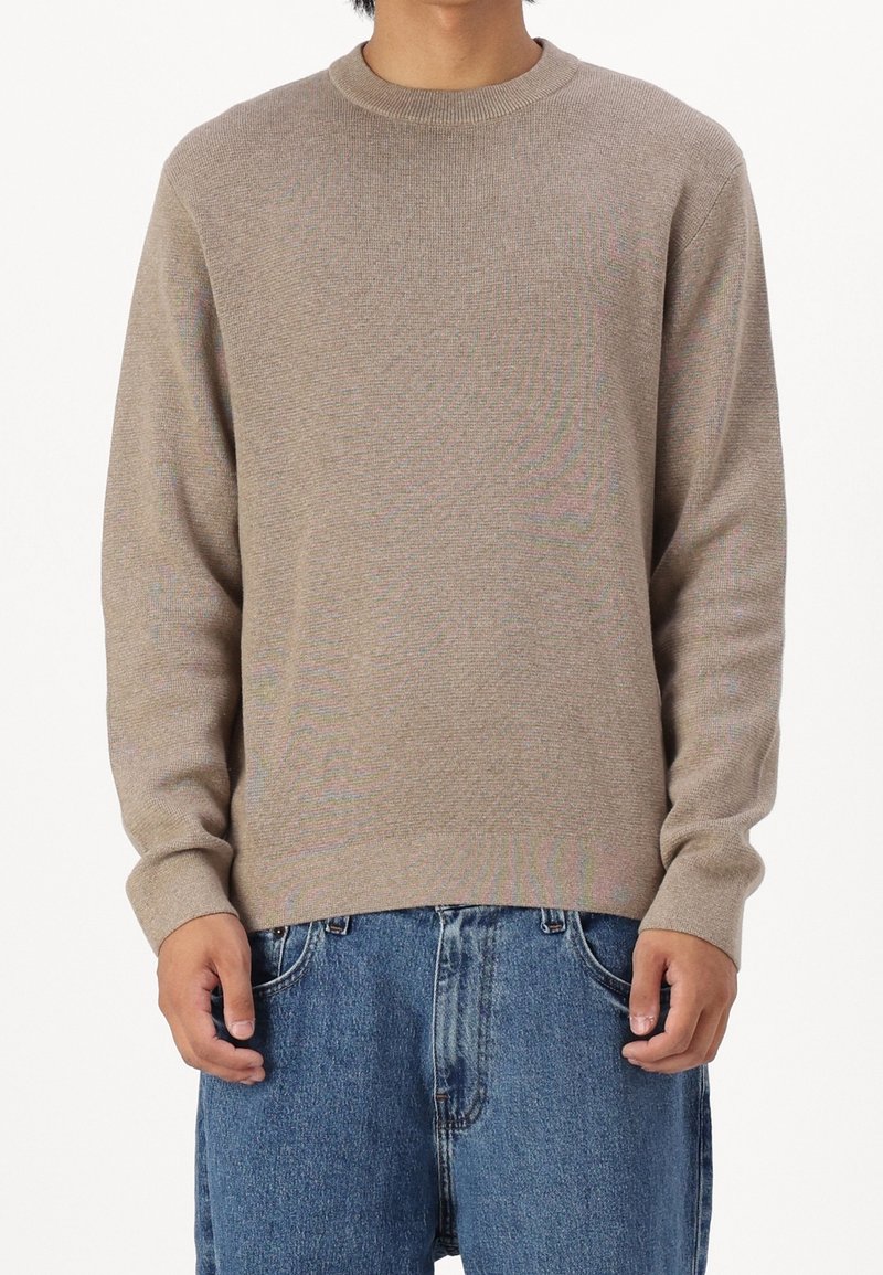 Homme portant un pull beige à col rond et un jean bleu en denim, debout, les mains détendues devant.