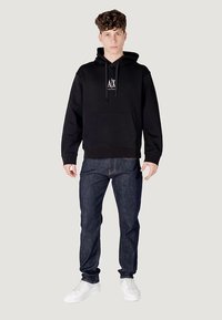 Fekete kapucnis pulóver első zsebbel és húzózsinórokkal, fehér logóval díszítve. Sötétkék farmernadrággal és fehér tornacipővel párosítva.