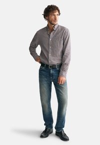 Camisa de cuadros en azul, blanco y marrón; vaqueros azules con un acabado desgastado; cinturón y zapatos negros. Pose de pie.