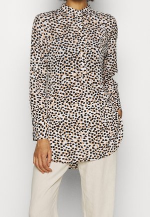 Chemise à manches longues avec motif de points irréguliers beige, noir et marron, associée à un pantalon beige clair, main dans la poche de la chemise.