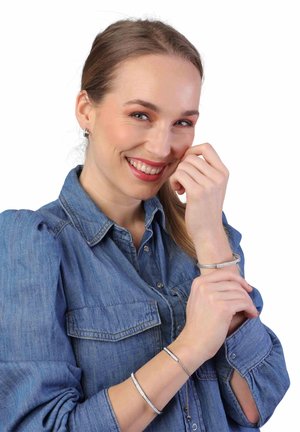 Zilveren metalen armbanden met verschillende texturen, gedragen om een pols. De persoon glimlacht en draagt een spijkerblouse met uitlopende mouwen.