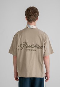 Beige rövid ujjú ing, hátulján sötétbarna felirattal: "Tilos" és "10119 Berlin". Színes mintás gallérral rendelkezik.