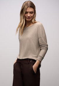 Beiger, leichter Pullover mit weitem Ausschnitt und langen Ärmeln, kombiniert mit dunkelbraunen Hosen mit seitlicher Tasche und Knopfdetail.