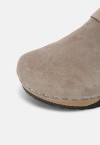 Gros plan sur l’avant d’une chaussure en daim taupe avec coutures visibles et semelle noire résistante sur fond blanc uni.