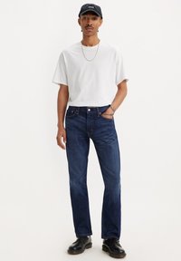 Camiseta blanca de cuello redondo, jeans azul oscuro, zapatos negros, cadena dorada y gorra negra; ajuste casual con un diseño minimalista y textura suave.