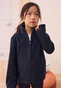 Giacca nera in pile con cerniera e collo alto, caratterizzata da una texture liscia e tasche laterali, abbinata a una maglietta bianca.