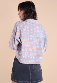 Femme de profil portant une veste à motifs bleu clair et corail avec une jupe en jean noire, devant un fond beige uni.