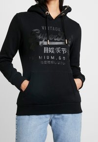 Felpa nera con cappuccio e tasca frontale, dotata di cordini. Presenta dettagli metallici. Caratterizzata da logo e testo stampati in colori a contrasto. Tessuto morbido.