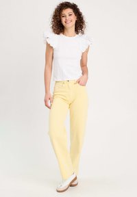 T-shirt blanc à manches volantées, assorti à un jean jaune taille haute. Modèle portant des baskets blanches avec des accents beige.
