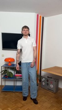 Noor mees tätoveeringutega, kandes valget polo särki ja heleid teksaseid, seisab toas, kus on värvilised seinavööd, oranž lamp, teler ja kohver.