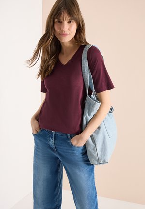 Femme portant un t-shirt bordeaux à col en V et un jean bleu, avec un sac à bandoulière bleu clair, debout les mains dans les poches.