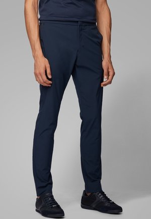 Mężczyzna w granatowych, dopasowanych spodniach i czarnych sneakersach, stojący na jednolitym szarym tle.