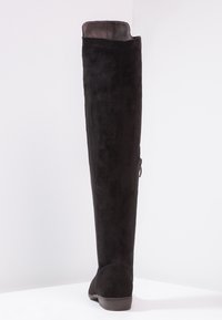 Bota de ante negra hasta la rodilla con un bajo tacón de madera y un cierre lateral, de pie contra un fondo blanco.