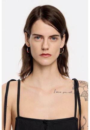Black spaghetti strap top with a lightly textured finish. Model wears colorful beaded hoop earrings. Prominent tattoos on the shoulder.