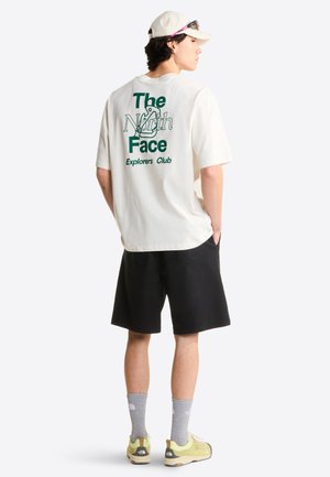 Persoon draagt een wit T-shirt met "The North Face Explorers Club"-opdruk, zwarte korte broek, grijze merkzokken, gele sneakers, witte pet en zonnebril op de pet.