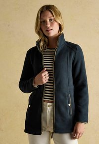 Tmavo modrá fleecová bunda s vysokým golierom, zipsom a zlatými akcentmi. Prúžkovaná košeľa pod ňou, svetlé nohavice, voľný strih.