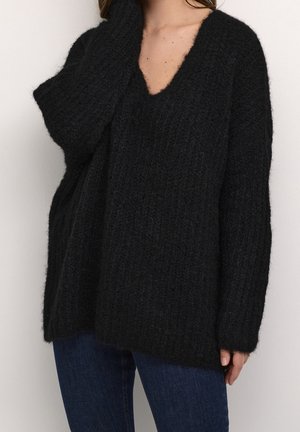 Femme portant un pull noir en laine à col en V ample avec des manches longues et un jean bleu, sur un fond uni.