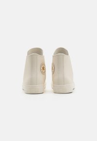 Beige hoge sneakers van glad materiaal, met een cirkelvormig logo op elke hiel en textuur rubberen zolen. Minimalistisch ontwerp.