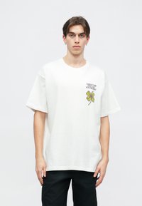 Biała koszulka z bawełny o swobodnym kroju, z krótkimi rękawami, z zielonym nadrukiem czterolistnej koniczyny oraz czarnym tekstem powyżej. Noszona z czarnymi spodniami.