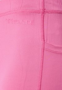 Zbliżenie różowego materiału z widocznym przeszyciem i wytłoczonym logo „hummel” w pobliżu bocznej kieszeni na obcisłej odzieży sportowej.