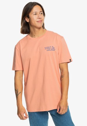 Peach-colored cotton t-shirt featuring a blue "Quik Silver" logo on the chest, short sleeves, and a relaxed fit, paired with blue jeans.