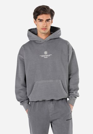 Grå sweatshirt med huva, framficka och vit text. Tyget verkar mjukt med en avslappnad passform. Modellen står mot en vit bakgrund.