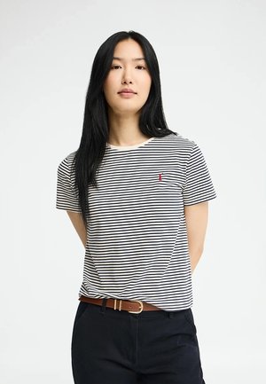 Jeune femme aux longs cheveux noirs portant un t-shirt à manches courtes rayé noir et blanc, un pantalon foncé et une ceinture marron, debout avec les mains derrière le dos.