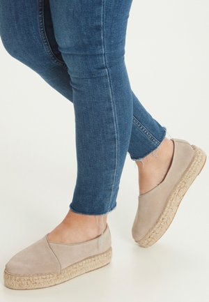 Fødder iført beige slip-on espadrillos med vævede såler, kombineret med blå skinny jeans med frynset kant.
