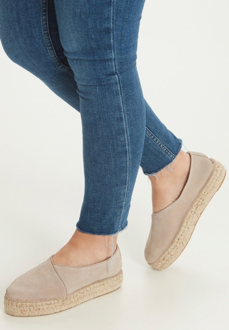 Pieds portant des espadrilles beige sans lacets avec des semelles tissées, associées à un jean skinny bleu aux ourlets effilochés.