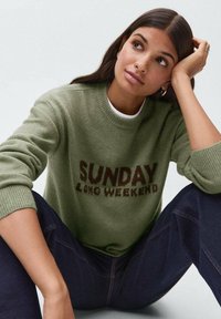 Svijetlozelena majica s natpisom "Nedjelja dugog vikenda" u tamnosmeđoj boji. Riblji rukavi i izrez, nosi se preko bijele majice, u kombinaciji s tamnim trapericama.