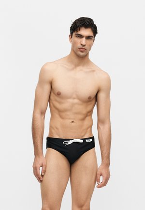 Lihaksikas mies, jolla on lyhyet tummat hiukset ja mustat uimahousut, joissa valkoinen kiristysnauha ja logo yksivärisellä vaalealla taustalla.