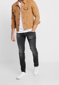En tan denimjacka med knappstängning, två bröstfickor, grå slitna jeans och vita sneakers. Tyget verkar mjukt och strukturerat.