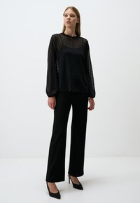 Schwarze Bluse mit Punkten, langen transparenten Ärmeln, rundem Halsausschnitt und lockerer Passform. Kombiniert mit schwarzen Hosen und hochhackigen Schuhen.