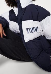 Person som bär en navy- och vit puffjacka med "TOMMY JEANS"-logotyp, tillsammans med svarta byxor, sittande mot en enkel bakgrund.