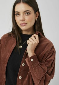 Chemise en velours côtelé marron à coupe décontractée, avec de gros boutons et une texture côtelée. Portée par-dessus un haut noir, accompagnée d'un collier floral.