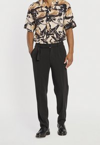 Beigen, mustan ja ruskean abstraktikuvioisen lyhythihaisen paidan yhdistelmä mustien räätälöityjen housujen ja mustien nyörikenkiin kanssa.