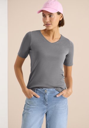 Femme portant un t-shirt gris à col en V et manches courtes, un pantalon rayé bleu avec les mains dans les poches, et une casquette de baseball rose, regardant sur le côté.
