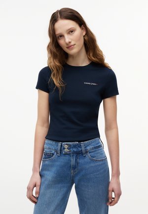 Young woman with wavy brown hair wearing a fitted navy "tommy jeans" t-shirt and high-waisted blue jeans with double buttons.