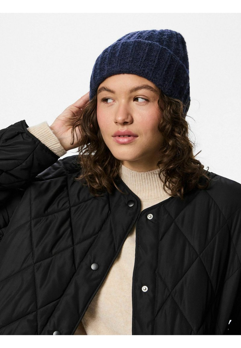 Stickad blå mössa med ribbad textur, svart vadderad jacka med diamantmönster och beige tröja. Handen är uppsträckt mot mössan.