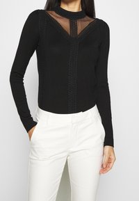 Svart långärmad bodysuit med en genomskinlig mesh-krage och detaljerad spetskant längs framsidan, parat med vita skräddarsydda byxor.