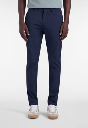 Mørkeblå skræddersyede bukser med en tætsiddende pasform. Fremstillet af glat stof, med bæltestropper, lynlås og sidelommer. Brugt med hvide sneakers.
