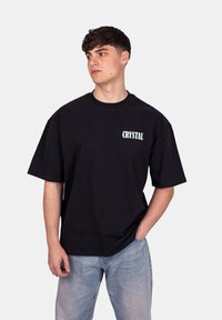 T-shirt noir oversize en coton. Équipé de manches courtes et d'un logo blanc "CRYSTAL" sur la poitrine. Porté avec un jean bleu clair.