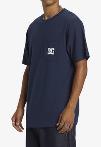 Námorně modré bavlněné tričko s krátkými rukávy, s kapsou na levé hrudi a bílým logem DC tištěným na kapse.