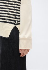 Κοντινό της χειρός ενός ατόμου δίπλα σε μια κρεμ πουκάμισο με μακριά μανίκια και κεντημένο μανσέτα, μαύρο πλεκτό γιλέκο με ρίγες και σκούρο παντελόνι.