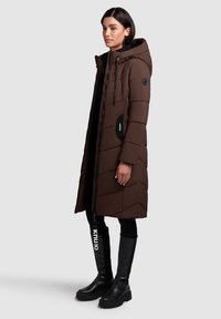 Palton puf maro lung cu glugă, având un model în zigzag, închidere cu fermoar și accente de buzunare laterale. Asortat cu cizme negre până la genunchi.