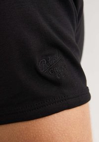 Tricou din bumbac negru cu un mic logo embosat pe mânecă, textură netedă și un design ajustat cu o margine curbată.