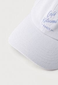 Casquette de baseball blanche avec texte brodé en bleu clair "Café Kitsuné Paris" sur le panneau avant, présentée sur une surface blanche.
