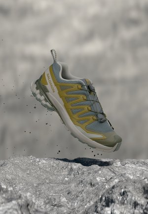 Trailrunning-schoen in grijze en gele tinten met een robuuste zool, zwevend boven rotsachtig terrein met kleine vallende stenen.